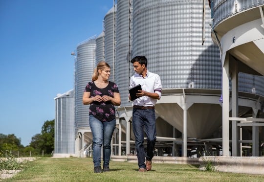 Two people walk by grain silos, representing a network of growers that enhances sales in the agricultural sector. Two people walk by grain silos, representing a network of growers that enhances sales in the agricultural sector.
